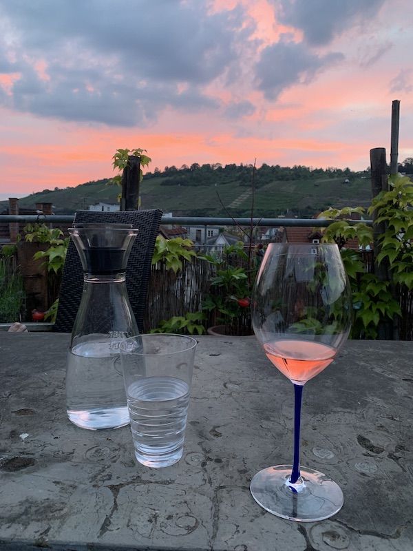 Abenddämmerung auf der Dachterrasse mit Blick in die Weinberge. Auf dem Steintisch steht eine Wasserkaraffe und ein Glas Roséwein. Der Wein hat die gleiche Farbe wie das Abendrot.