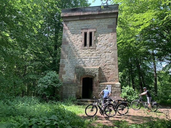 Zwei Fahrradfahrer vor einem alten Turm mitten im Wald. Die Sonne scheint durch die Bäume und es ist eine friedliche Stimmung.